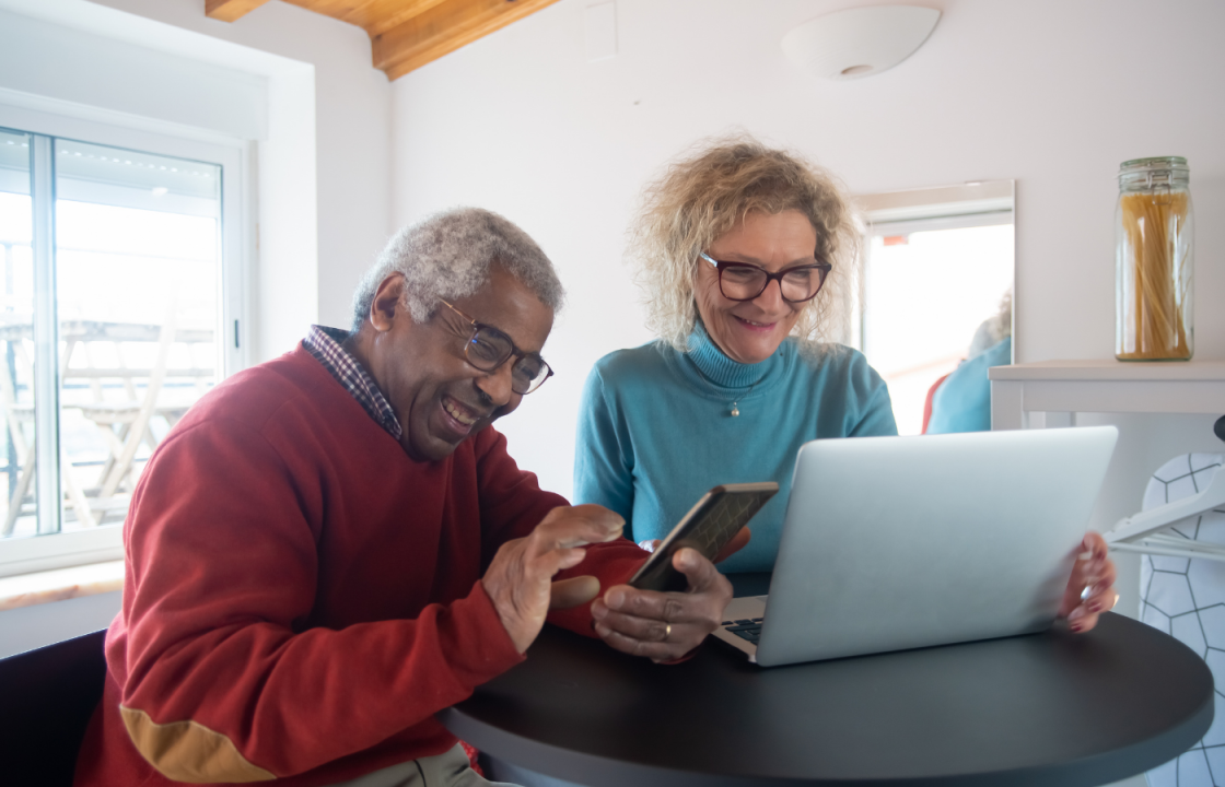 Black older male and white older female sitting at table smiling as they look at phone and computer