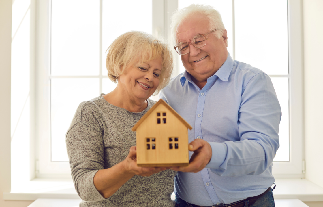 white older adults standing close smiling looking at wood home model in their hands